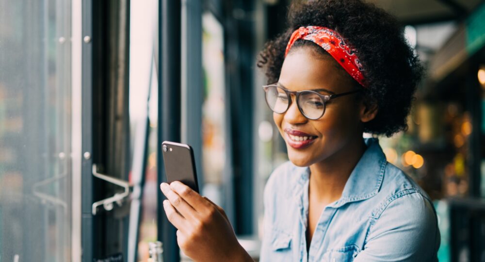Attractive young African woman smiling and reading texts on a cellphone while sitting alone at a counter in a cafe enjoying a meal