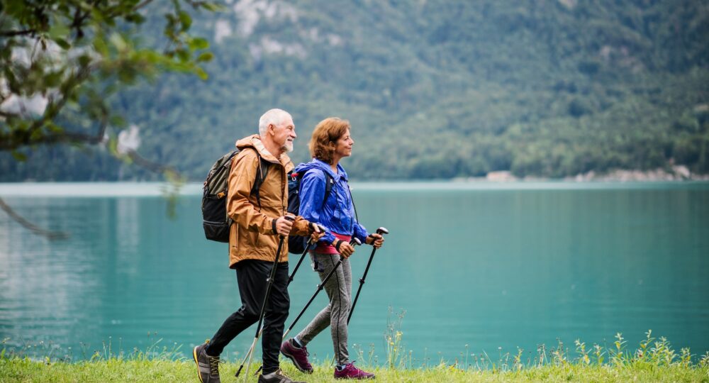 A side view of senior pensioner couple hiking by lake in nature, talking. Copy space.