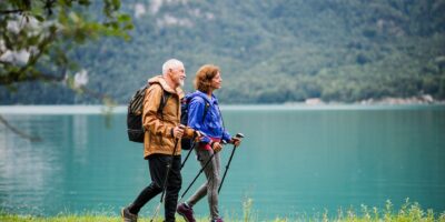 A side view of senior pensioner couple hiking by lake in nature, talking. Copy space.
